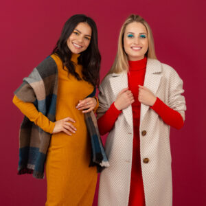 two smiling attractive stylish women in autumn winter fashion dress and coat posing isolated on red studio background